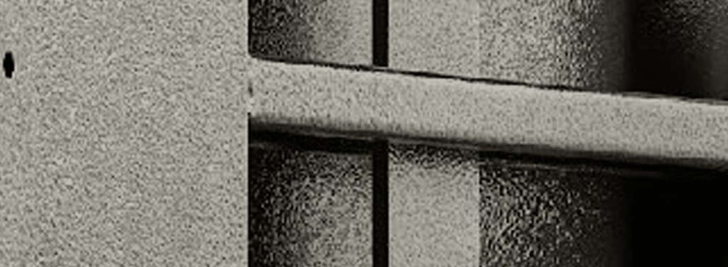 Close-up of a textured, metallic jail or prison cell door with vertical and horizontal bars, seen in black and white.