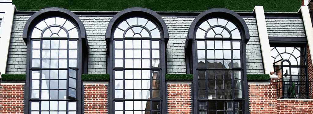 Three large arched windows with black frames set in a brick building with a slate-tiled roof and green artificial grass above the windows.