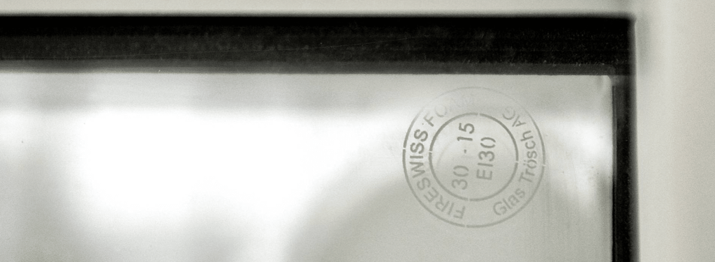 Close-up of a windowpane with a circular manufacturer stamp reading “SWISS-F” and “Glas Trösch AG” along with numbers and letters, photographed through the glass.