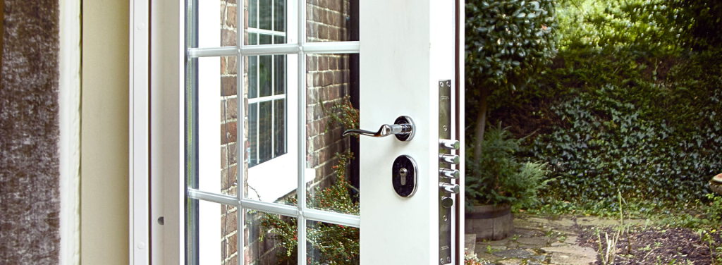 A white door with glass panes is slightly open, revealing a lush green garden outside. Sunlight filters through the open door, and brick walls and plants are visible in the background.