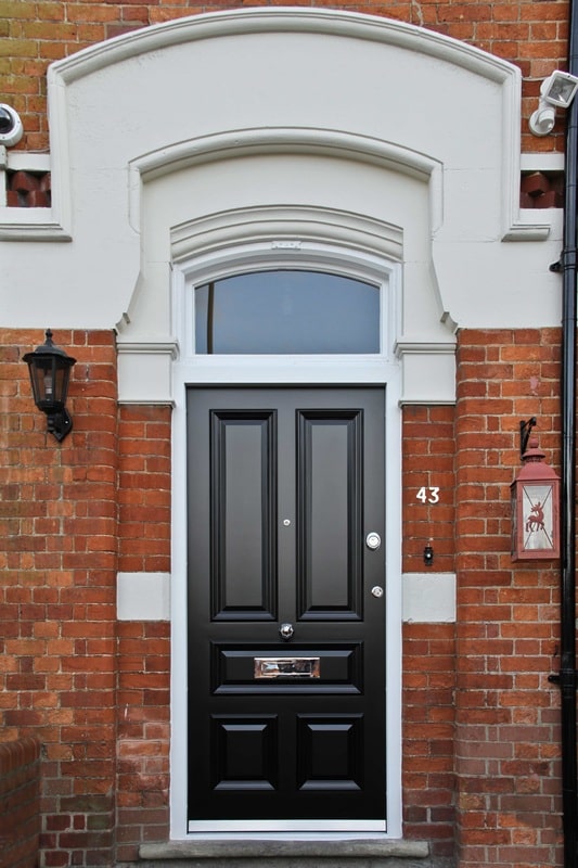 A black front door with decorative panels set in a red brick wall, featuring a white arched frame, transom window above, and house number 43 to the right. A lantern-style light and a red mailbox are mounted on the wall.