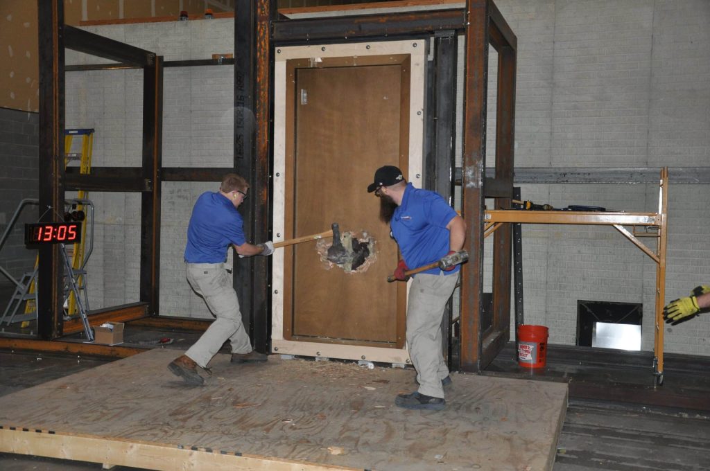 Two individuals using hammers to put a hole in a wooden door.