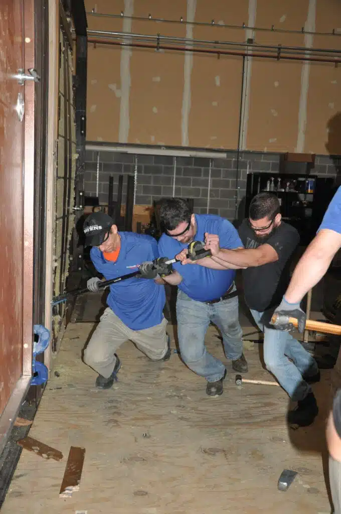 Three men working together using a pry bar to force open a large door in an indoor training facility, with protective gloves and eyewear.