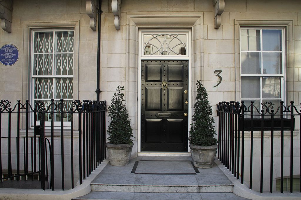 A stone townhouse entrance with a black door marked number 3, flanked by two potted topiary trees and black metal railings; white-framed windows and a blue plaque are visible on the left.