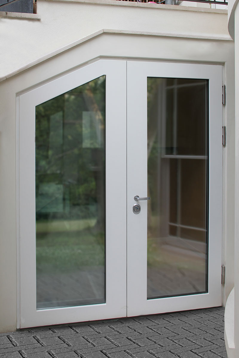 A modern white door with large angled glass panels, set against a white exterior wall. The door is situated beneath a staircase, with reflections of greenery and outdoor paving visible through the glass.