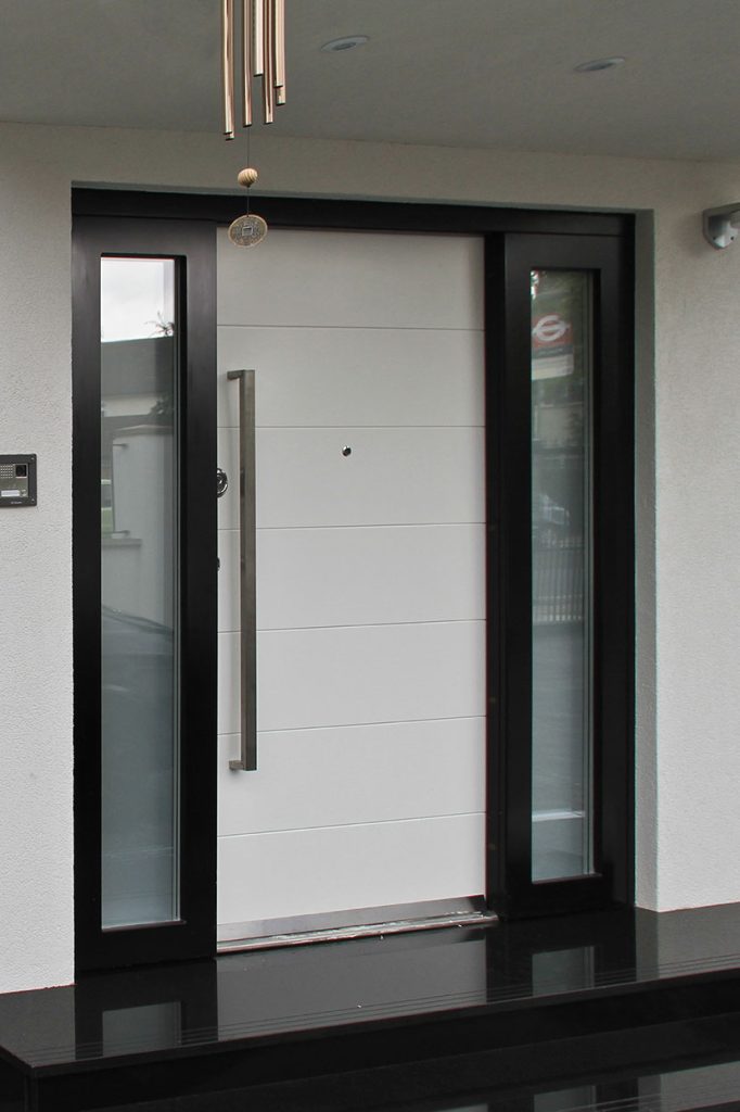 A modern front door with white horizontal panels, a tall silver handle, and side glass panels. The door is framed in black, with a doorbell and camera on the left wall and a small chime hanging above.