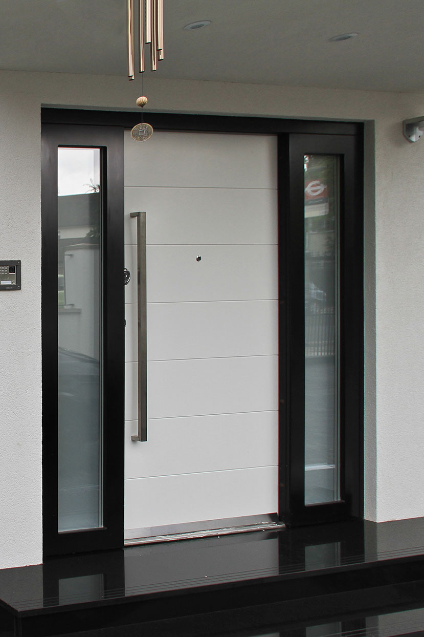 A modern front door with white horizontal panels, a tall silver handle, and side glass panels. The door is framed in black, with a doorbell and camera on the left wall and a small chime hanging above.