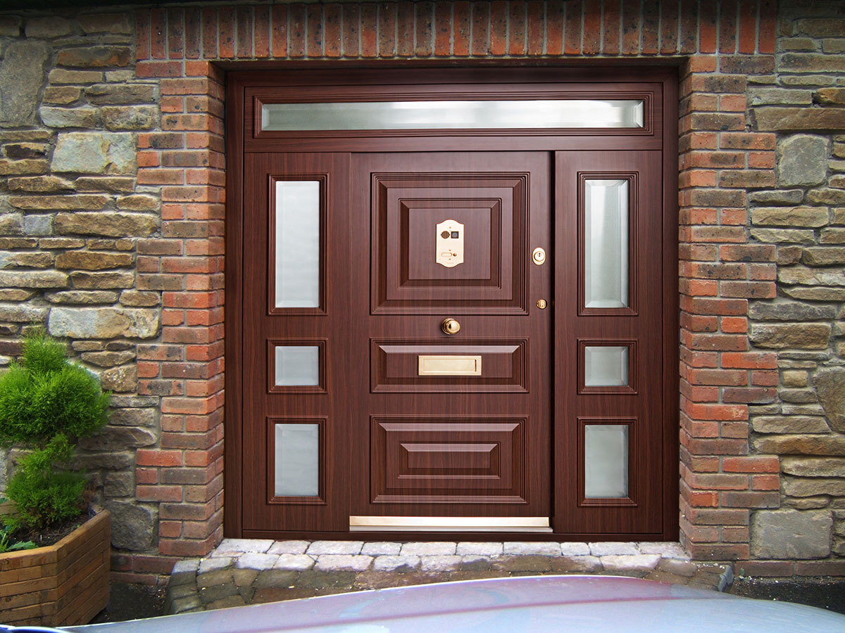 A wooden front door with geometric panel designs, surrounded by brick and stone walls. The door features frosted glass panels, a brass mail slot, door knocker, and handles, with a small potted plant nearby.