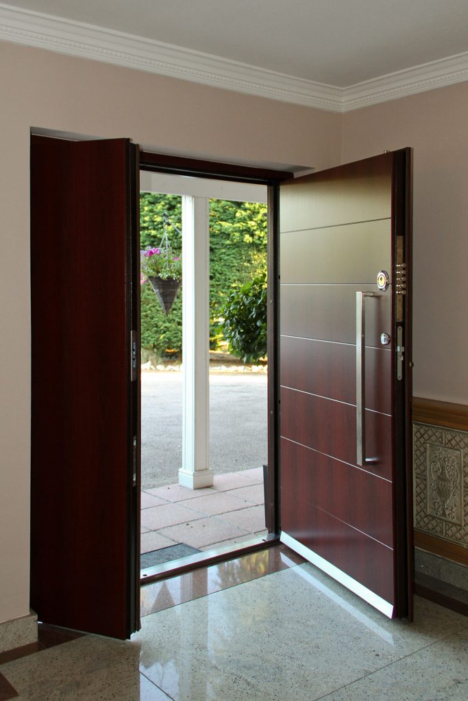 A modern, dark wooden front door is open, revealing a tiled entryway and an outdoor area with greenery and a hanging flower basket. The interior has beige walls and a glossy marble floor.