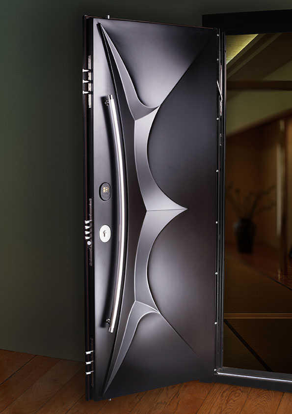 A modern, metallic black door with a sculpted geometric design and a long vertical handle is partially open, revealing a dimly lit room with wooden flooring and blurred decor in the background.