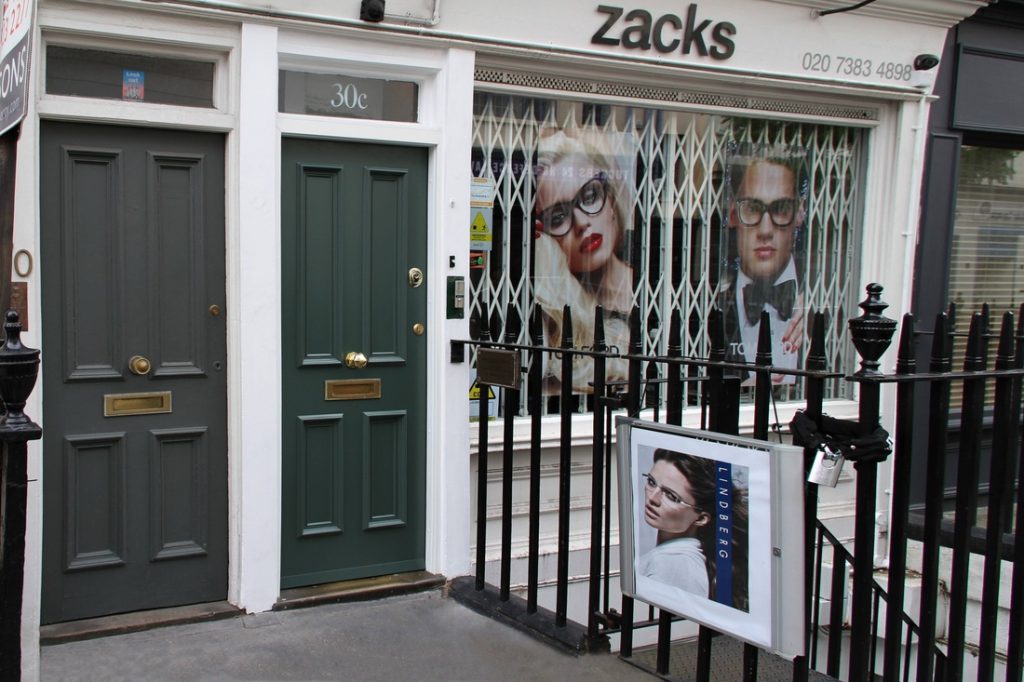 A shopfront with the sign zacks above, metal security bars on the window, and large posters of people wearing glasses. To the left are two green doors labeled 30 and 30c, and a poster featuring a woman is attached to the railing.
