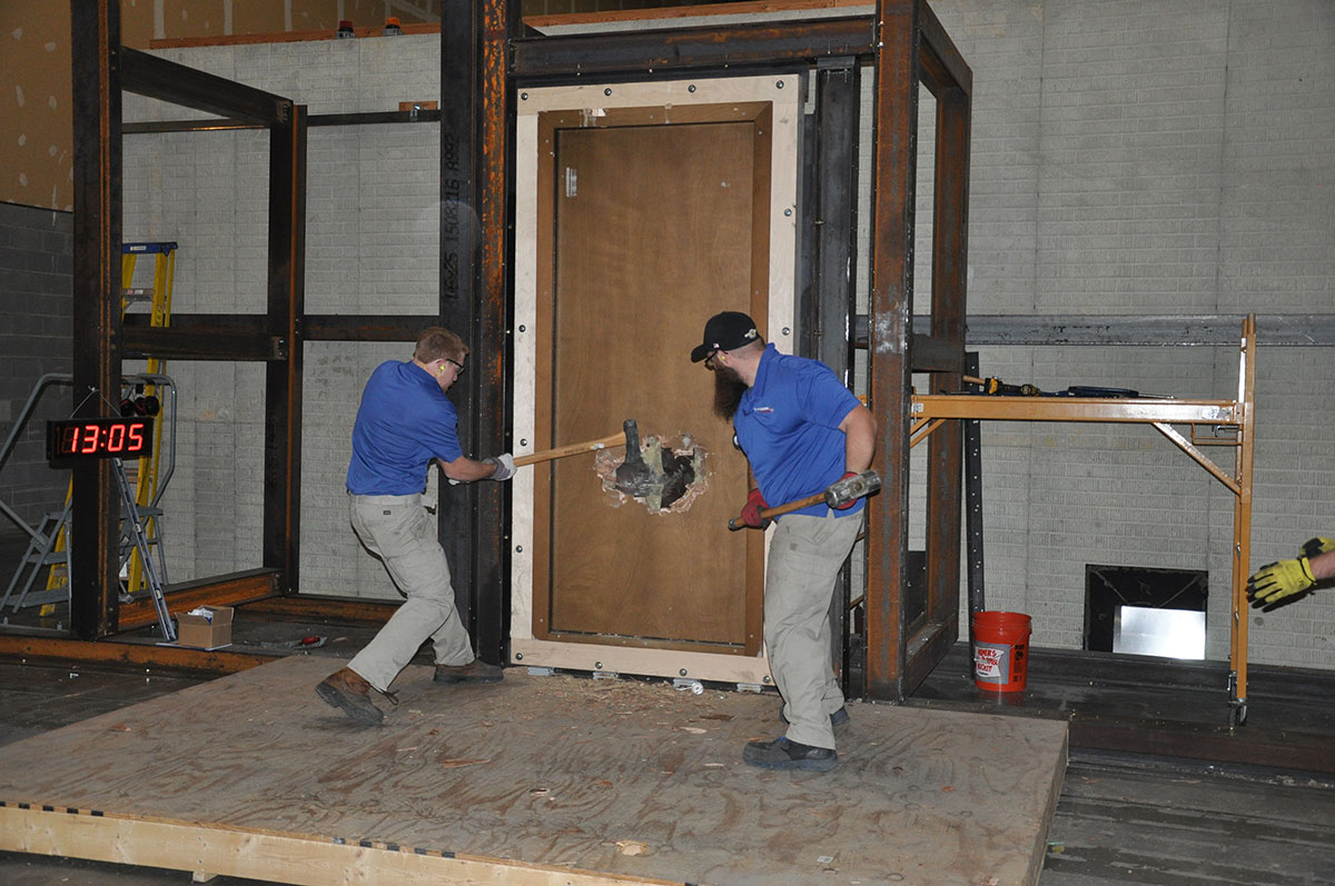 Two men in blue shirts and khaki pants use sledgehammers to break through a large wooden door, creating a hole in the center. Construction materials and a digital timer are visible in the background.