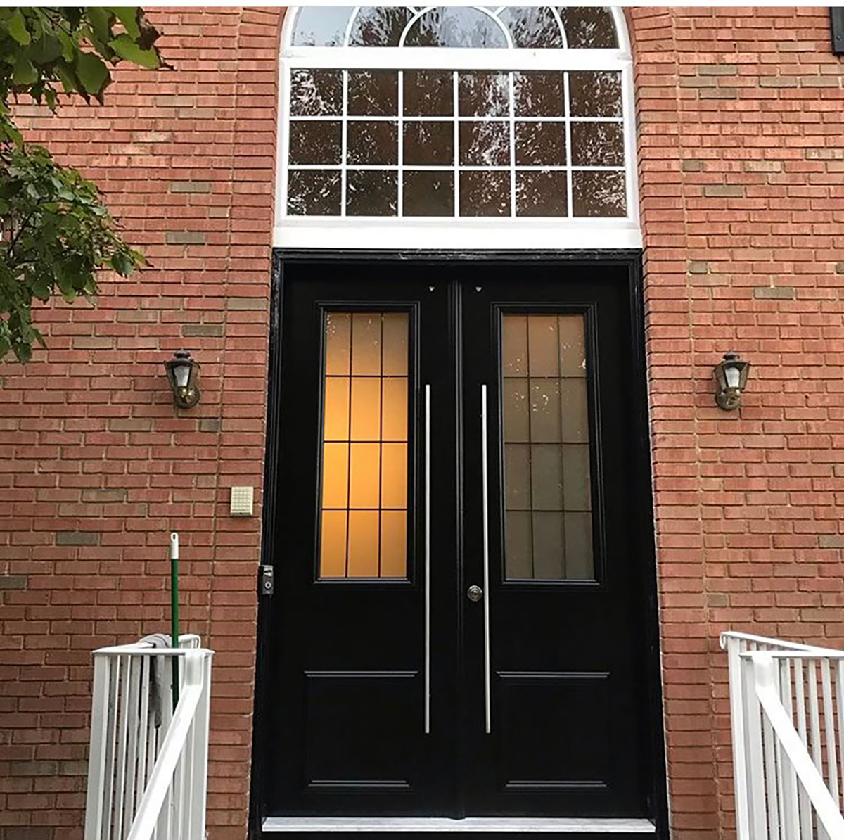 A pair of black double doors with vertical glass panels and long handles, set in a red brick wall with a large arched window above. Two wall lamps and white railings frame the entrance. A broom leans against one railing.