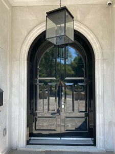 A glossy black double door with an arched frame and a large, modern glass lantern hanging above, set in a light stone exterior. Trees and sky are reflected in the door’s surface.