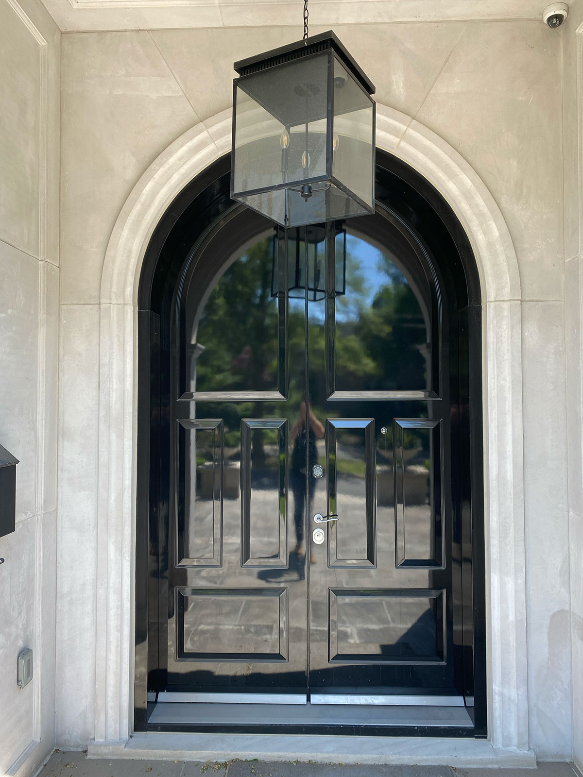 A glossy black double door with an arched frame and a large, modern glass lantern hanging above, set in a light stone exterior. Trees and sky are reflected in the door’s surface.