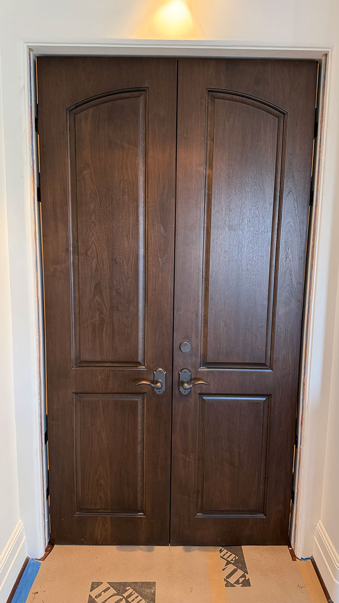 A pair of closed, dark wooden double doors with arched panels, bronze handles, and white trim set in a white wall. The floor in front of the doors is partially covered with protective material.