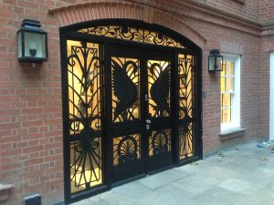 Black wrought iron double doors with intricate designs, including large crane or stork silhouettes, set in a red brick building with two wall lanterns and a window on the right. Warm light glows from inside.
