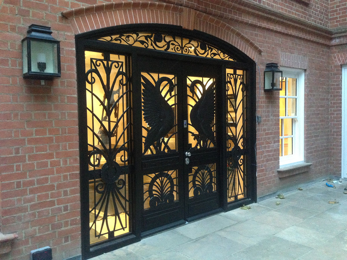 Black wrought iron double doors with intricate designs, including large crane or stork silhouettes, set in a red brick building with two wall lanterns and a window on the right. Warm light glows from inside.