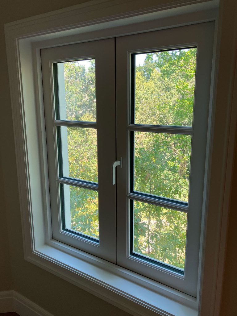 A white-framed double window with a handle at the center, looking out onto leafy green trees and sunlight outside. The window has four glass panes on each side.