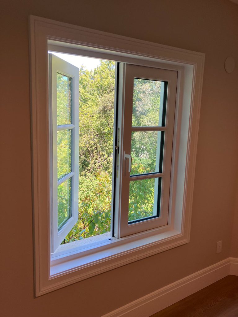 A white-framed window is open inward, letting in sunlight and revealing green trees and foliage outside. The room inside has beige walls and a wooden floor.