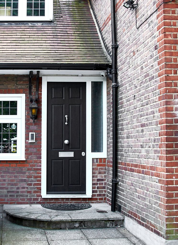 A black front door with a silver mail slot and door knocker, set in a red brick house with white trim; a doormat and outdoor light are beside the entrance.