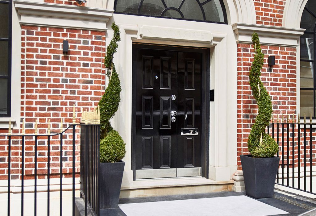 A black door with silver hardware is set in a red brick building with arched windows. Two spiral-shaped topiary bushes in square planters stand on either side of the entrance, which is framed by light stone.