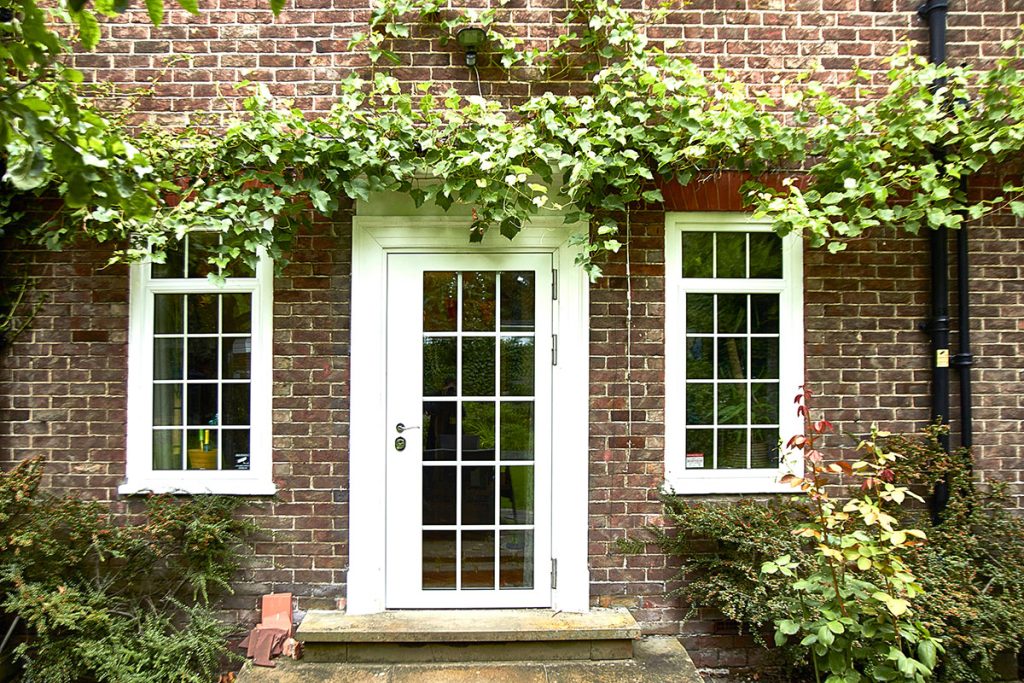 A white-framed glass door is centered between two white-framed windows on a brick wall, with green vines growing above and around the door and windows. Bushes and plants line the base of the building.