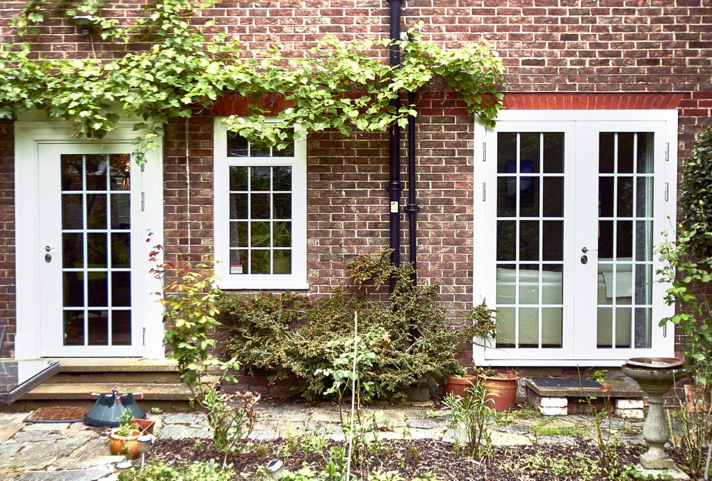 A brick house exterior with two white French doors and a window in between, surrounded by green vines and plants. The patio area has various potted plants and a birdbath on the right.