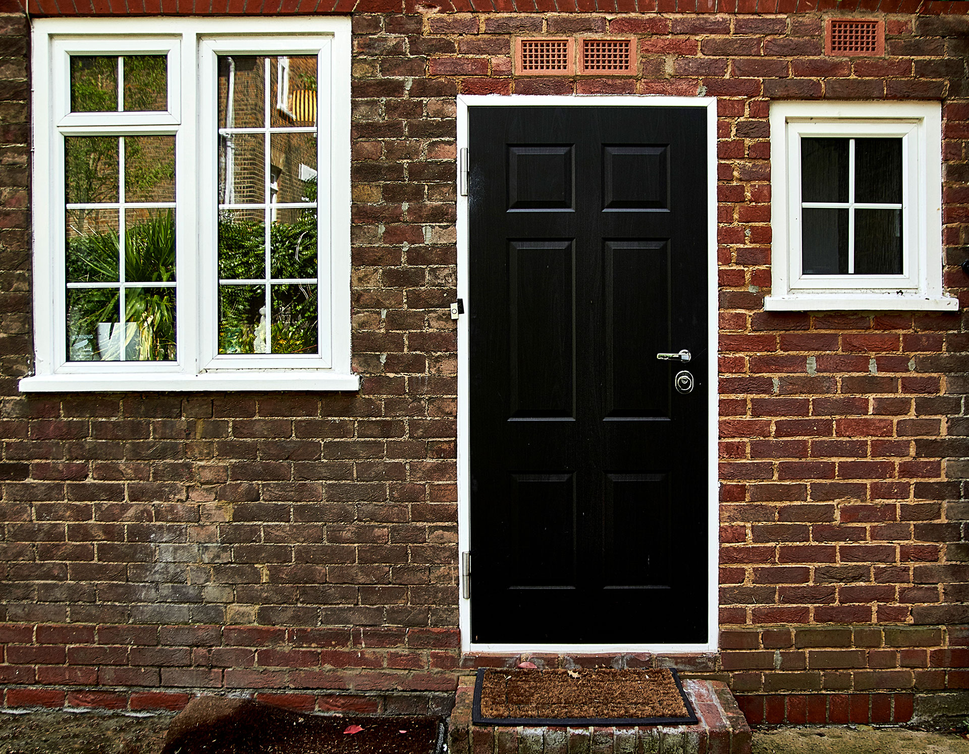 A black front door with a silver handle is centered in a brick wall, flanked by a large white-framed window on the left and a small square white-framed window on the right. A brown doormat sits in front of the door.