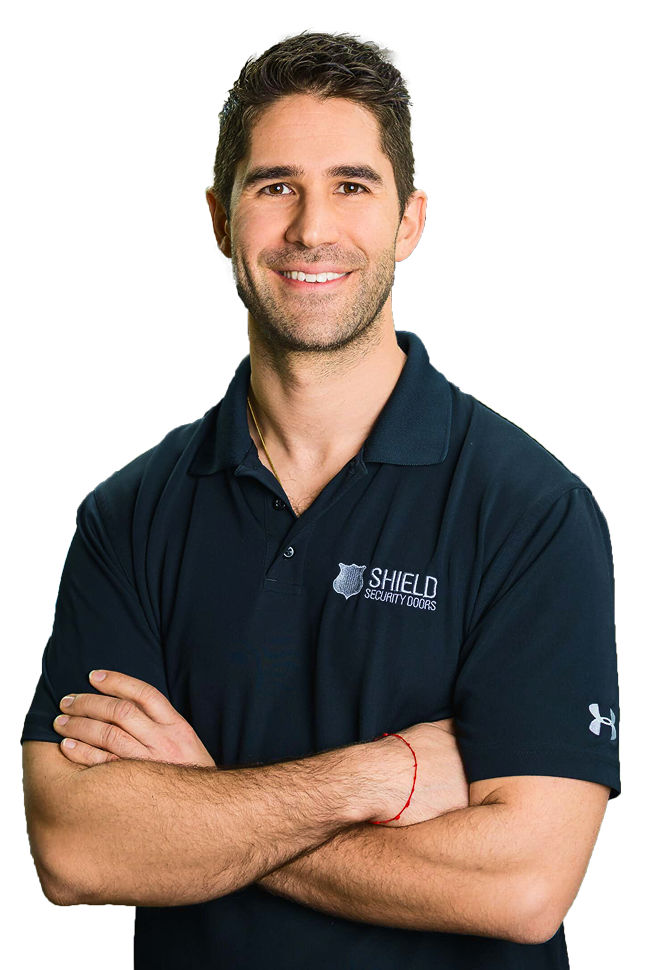 A man with short brown hair and a beard smiles while standing with arms crossed, wearing a dark blue SHIELD Security Doors polo shirt and a red string bracelet, against a plain white background.