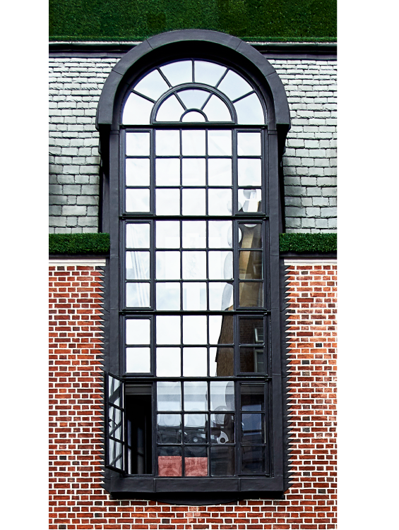 Tall, arched window with black grid framing set in a red brick wall; one lower pane is open. The window reflects the sky and nearby buildings. Gray roof shingles and green trim are visible above the window.