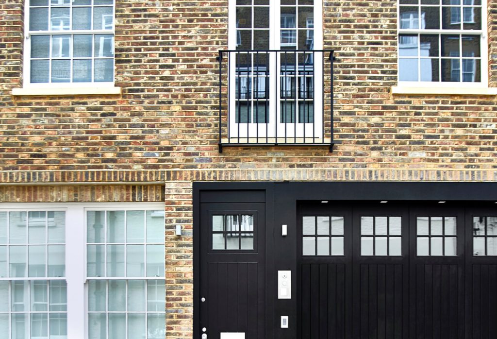 A modern brick house facade with large white-paned windows, a black front door, and a matching black garage door. There is a small balcony with a metal railing above the entrance.